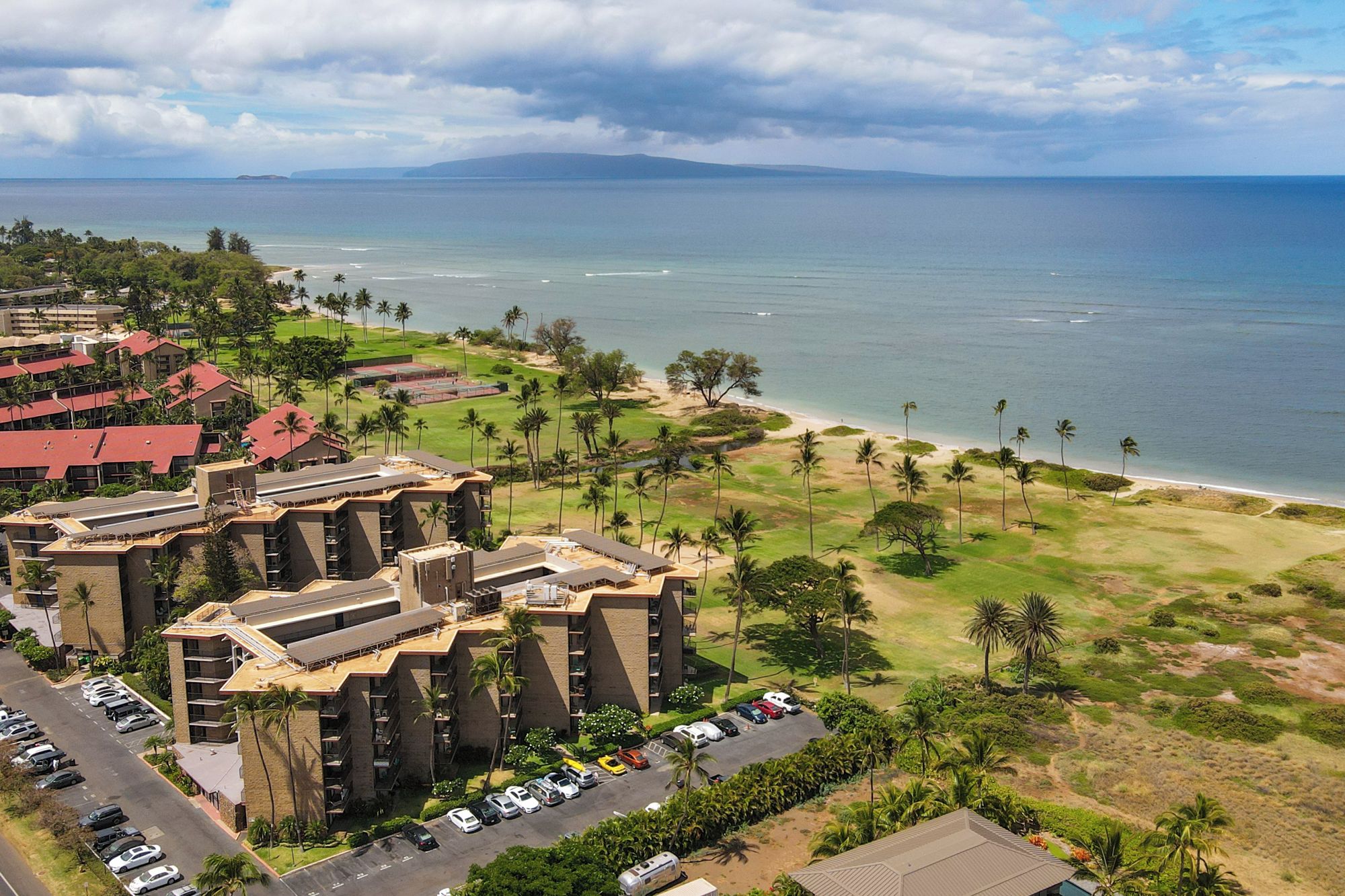 A coastal hotel with multiple buildings, a parking lot, and a beach area.