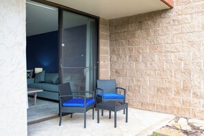 A balcony with two wicker chairs and a table, a sliding glass door leading to an interior room with a sofa and a lamp. - Oceanfront Kihei Condo | Ground Floor, Pool, Hot Tub & AC (Kihei)
