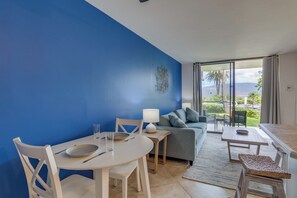 A modern living room with a blue accent wall, a sofa, a dining table with chairs, and a view of the outdoors. - Oceanfront Kihei Condo | Ground Floor, Pool, Hot Tub & AC (Kihei)