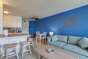 A modern living space with a blue accent wall, a dining area, and a kitchen with white cabinets. - Oceanfront Kihei Condo | Ground Floor, Pool, Hot Tub & AC (Kihei)
