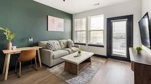 Uma sala de estar moderna com sofá, mesa de centro e escrivaninha com cadeira. Há janelas com persianas, uma planta e um quadro na parede.