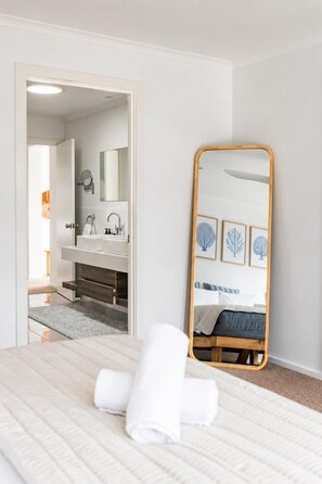 A bedroom with a bed, a large mirror, a bathroom visible through an open door, and a neatly made bed with a rolled towel. - Salt Bush | Swim Spa | Close to Everything (Bicheno)