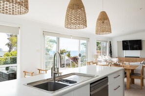 A modern kitchen with a large island, stainless steel sink, and overhead wicker pendant lights. - Salt Bush | Swim Spa | Close to Everything (Bicheno)