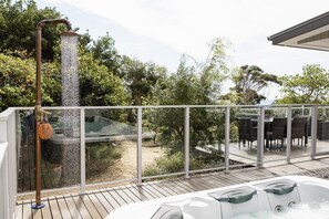 Uma banheira de hidromassagem em um deck de madeira com guarda-corpo de vidro, com vista para um jardim com árvores e um espaço de varanda.