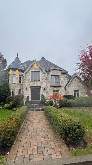 A stone house with a central entrance, a covered porch, and a brick pathway leading to the door. - Luxury Estate Home | Cinema & Sauna (Candiac)