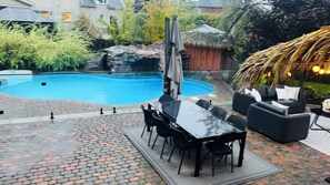 Piscina com cobertura de vidro, rodeada por um terraço com piso de tijolo e mobiliário de exterior.