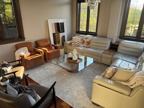 Ein Wohnzimmer mit einer Couch, Sesseln, einem Couchtisch und einem großen Fenster mit Blick auf Bäume.