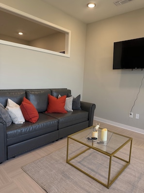 Un salon avec un canapé en cuir foncé, une table basse en verre, un téléviseur à écran plat fixé au mur et un luminaire encastré dans le plafond.