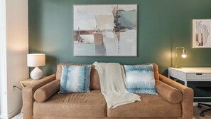 A brown sofa with a blanket and patterned cushions, a white lamp, and a desk with a lamp. - Landing | Chic 1BD, Pool, Courtyard (Orlando)
