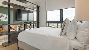A modern hotel room with a large bed, a flat-screen TV mounted on a wall, a view of the cityscape through large windows, and a balcony with a sofa. - Landing | Stunning Studio, Rooftop Lounge (Orlando)