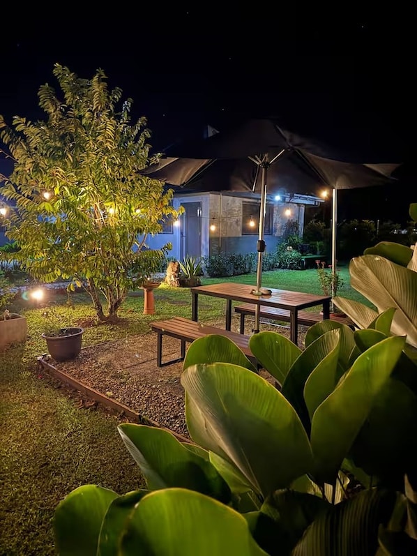 An outdoor patio area with a table and chairs, surrounded by greenery and a building in the background.