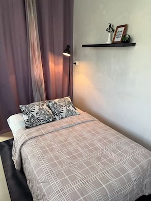 A bedroom with a bed, a bedside lamp, a shelf with decorative items, and a window with curtains. - Garden Guesthouse w/ Kitchen, AC, Starlink & Bikes – Pacific Harbour, Fiji (Pacific Harbour)