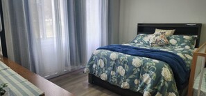 Quarto com cama, cortinas, piso de madeira e vista pela janela.