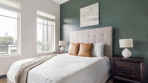 A bedroom with a large bed, two bedside tables, a window with blinds, and a city view. - Landing | Beautiful 1BD, Gym, Courtyard (Atlanta)