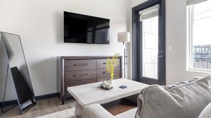 A modern living room with a flat-screen TV mounted on the wall, a dark wooden dresser with drawers, a table with a vase of flowers, and a large window with blinds. - Landing | Beautiful 1BD, Gym, Courtyard (Atlanta)