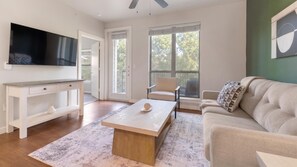 A living room with a flat-screen TV, a wooden coffee table, a beige sofa, and a large window with a view of trees. - Landing | Incredible 1BD, Gym, Pool (Austin)