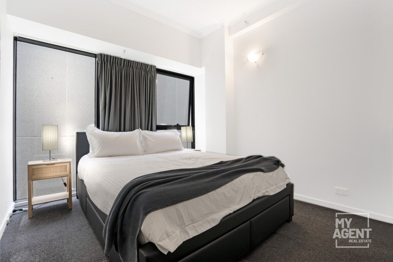 A modern bedroom with a large bed, a nightstand, a lamp, and a view of the outside through a window with curtains.