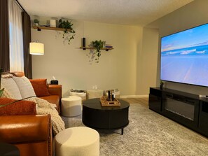 Un salon comprenant un canapé, un pouf, une table basse, un téléviseur et des étagères ornées de plantes et de livres.