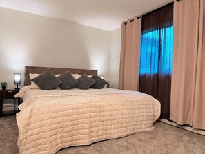Ein Schlafzimmer mit einem großen Bett, einem Nachttisch, einer Lampe und einem Fenster mit Vorhängen.