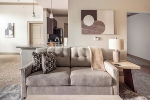 A modern living room with a grey sofa, a coffee table, and a lamp. There is a kitchen area in the background with a refrigerator and a sink. - Landing | Stunning 1BD, Gym (Destin)