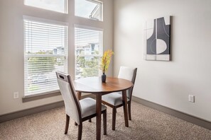A dining area with a round table, two chairs, a vase with yellow flowers, and a modern abstract painting on the wall. - Landing | Stunning 1BD, Gym (Destin)