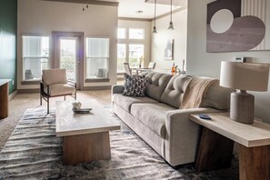 Una sala de estar con un sofá, una mesa de centro y una lámpara. Hay ventanas con persianas, una silla y un cuadro en la pared.