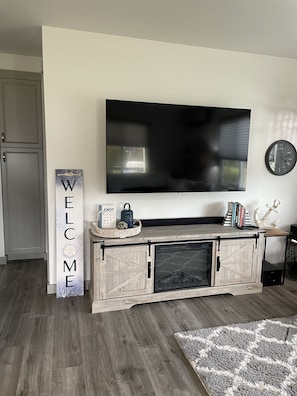 A living room with a mounted TV, a wooden console table with decorative items, a welcome sign, and a clock on the wall. - Caspers Cozy Cottage- Whidbey Island getaway (Clinton)