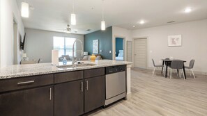 A modern kitchen with dark cabinets, a granite countertop, and a dishwasher. There is a dining area with a table and chairs. - Landing | Stunning 2BD, Rooftop Lounge, Clubhouse (Fort Worth)
