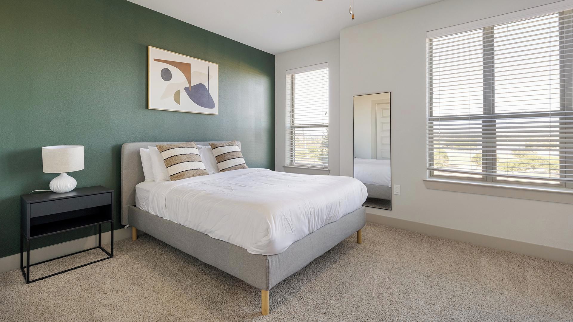 A bedroom with a bed, a bedside table, a lamp, a painting, and a window with blinds.