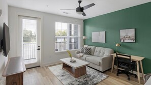 A modern living room with a sofa, coffee table, and a desk with a chair. There are two windows with blinds, a ceiling fan, and artwork on the wall. - Landing | Spectacular 1BD, Gym, Clubhouse (Huntsville)
