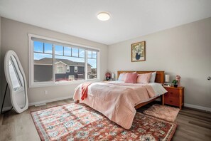 Een slaapkamer met een groot raam, een bed met een roze deken, een houten nachtkastje en een klein nachtkastje met een lamp.