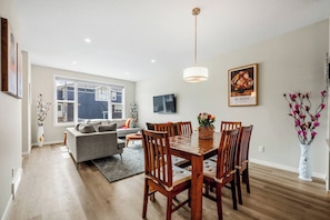 A modern living room with a dining area, a sofa, a TV, and a large window. - Corner Glen Retreat | 6BR | Sleeps 12 - Holiday Home (Calgary)
