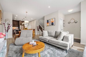 A modern living room with a grey sofa, a round wooden coffee table, and a dining area in the background. - Corner Glen Retreat | 6BR | Sleeps 12 - Holiday Home (Calgary)