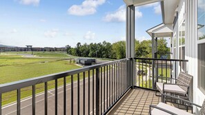 A balcony with a view of a road and greenery, a covered outdoor seating area, and a building with large windows. - Landing | Modern 1BD, Yoga Studio, Gym (Huntsville)