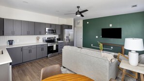A modern kitchen with grey cabinets, a microwave, stove, and refrigerator. There is a grey sofa, a wooden dining table, and a white lamp. A flat-screen TV is mounted on a green wall. - Landing | Stunning 2BD, Clubhouse, Gym (Huntsville)