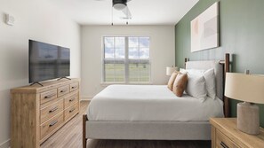 A bedroom with a bed, a TV on a wooden dresser, a window with blinds, and a ceiling fan. - Landing | Modern 1BD, Game Room, Gym (Huntsville)