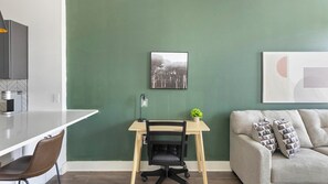 A modern living space with a green accent wall, a wooden desk with a chair, a grey sofa with patterned cushions, and a framed picture above the desk. - Landing | Modern 1BD, Game Room, Gym (Huntsville)