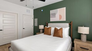 A hotel room with a bed, two bedside tables, a wall-mounted lamp, and a door. - Landing | Amazing 2BD, Game Room, Gym (Huntsville)