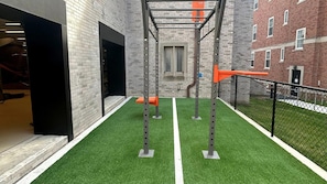 A gym area with artificial turf, exercise equipment, and a brick building in the background. - Landing | Incredible 1BD, Pool (West Hartford)