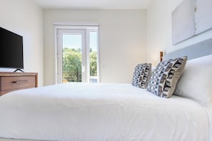 A bedroom with a large bed, a television on a wooden dresser, and a balcony with a view of trees. - Landing | Incredible 1BD, Pool (West Hartford)
