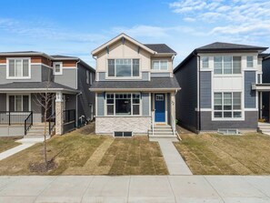 A two-story residential building with a stone facade, a blue front door, and a small front yard. - Corner Glen Retreat | 4BR Upper | Sleeps 8 - Holiday Home (Calgary)