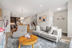 Ein modernes Wohnzimmer mit einer grauen Couch, einem runden Holztisch und einem Essbereich im Hintergrund.