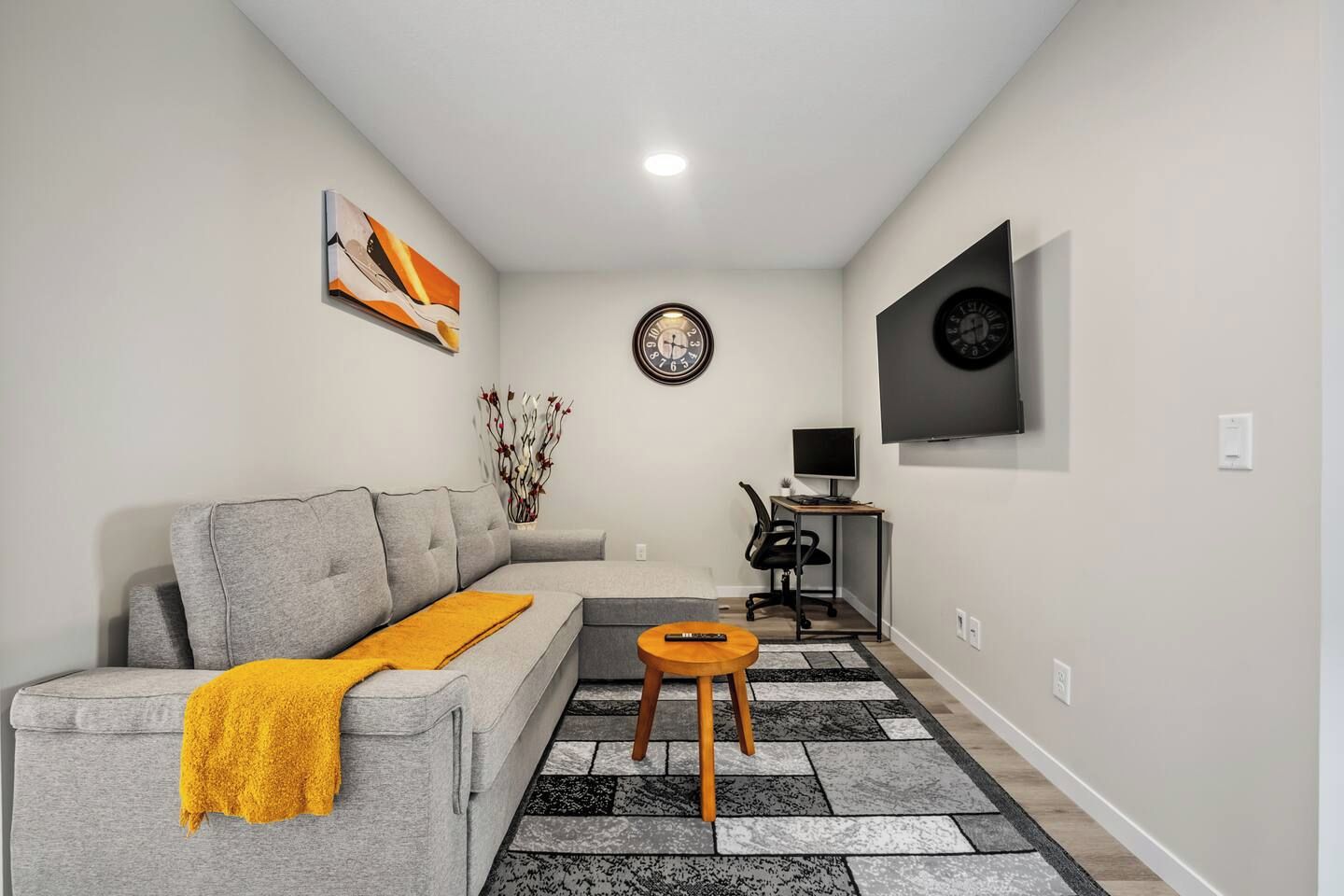 A modern living room with a grey sofa, a yellow throw blanket, a round wooden side table, a black and white patterned rug, a wall-mounted clock, a flat-screen TV, and a decorative vase.