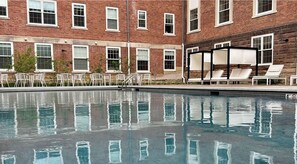 A swimming pool with a reflection of a brick building and white chairs. - Landing | Modern 1BD, Pool (West Hartford)