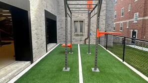 A gym area with artificial turf, exercise equipment, and a brick building in the background. - Landing | Modern 1BD, Pool (West Hartford)