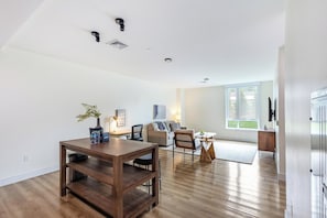 A spacious living room with a wooden dining table, a sofa, and a TV mounted on the wall. - Landing | Modern 1BD, Pool (West Hartford)
