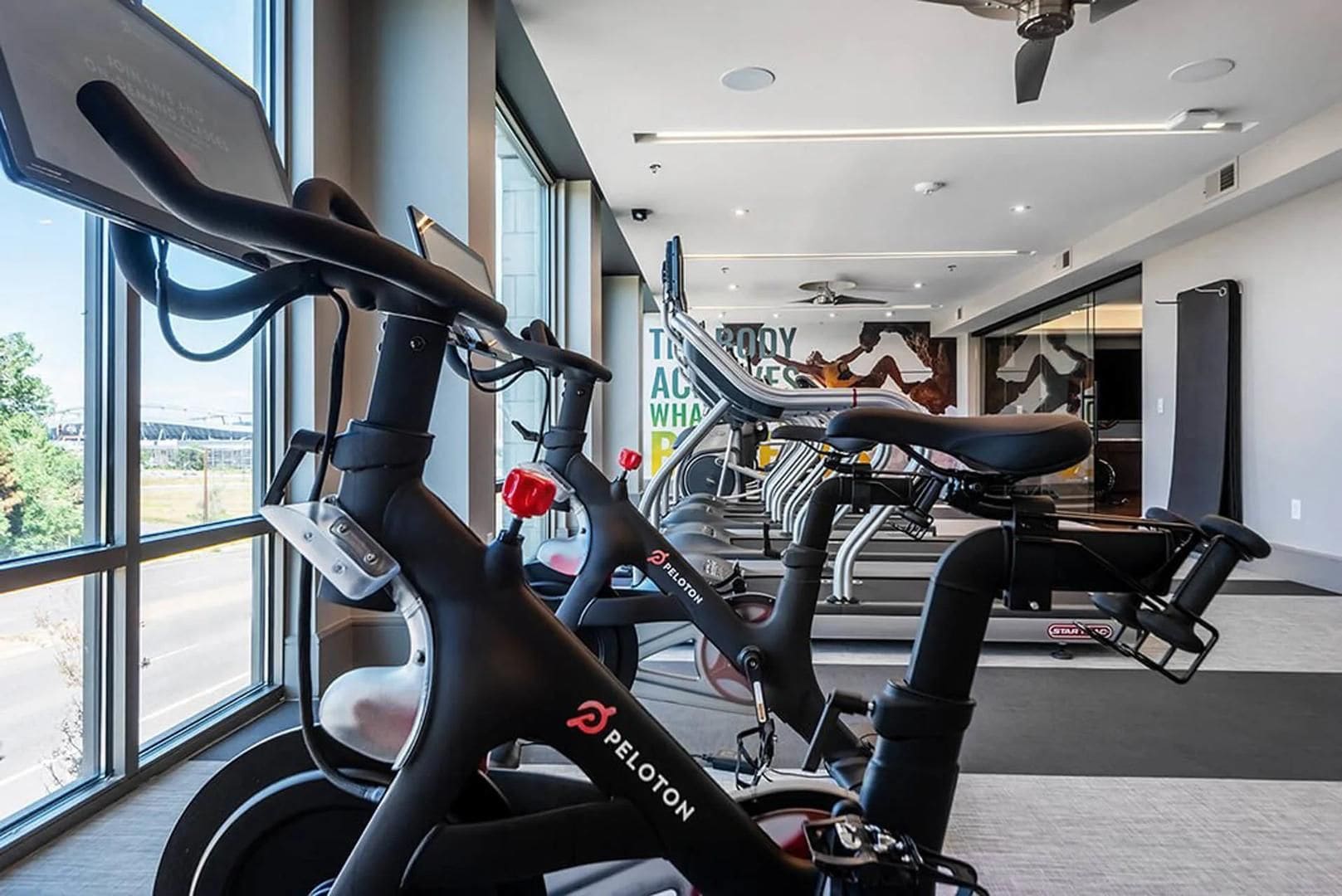 A gym with stationary bikes, large windows, and a ceiling fan.