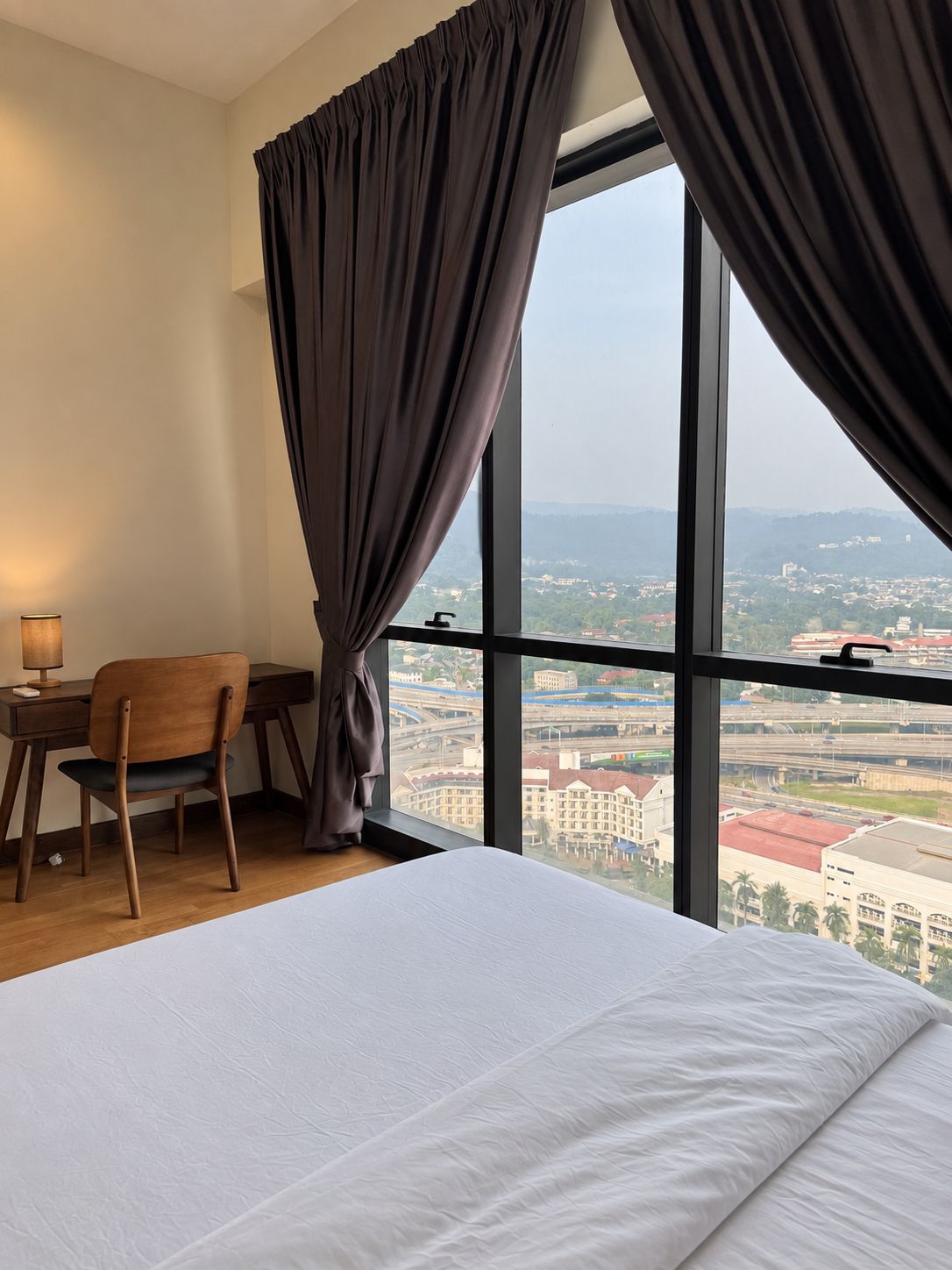 Ein Hotelzimmer mit einem großen Bett, einem Schreibtisch und einem Stuhl. Durch ein großes Fenster ist ein Blick auf die Stadt möglich.