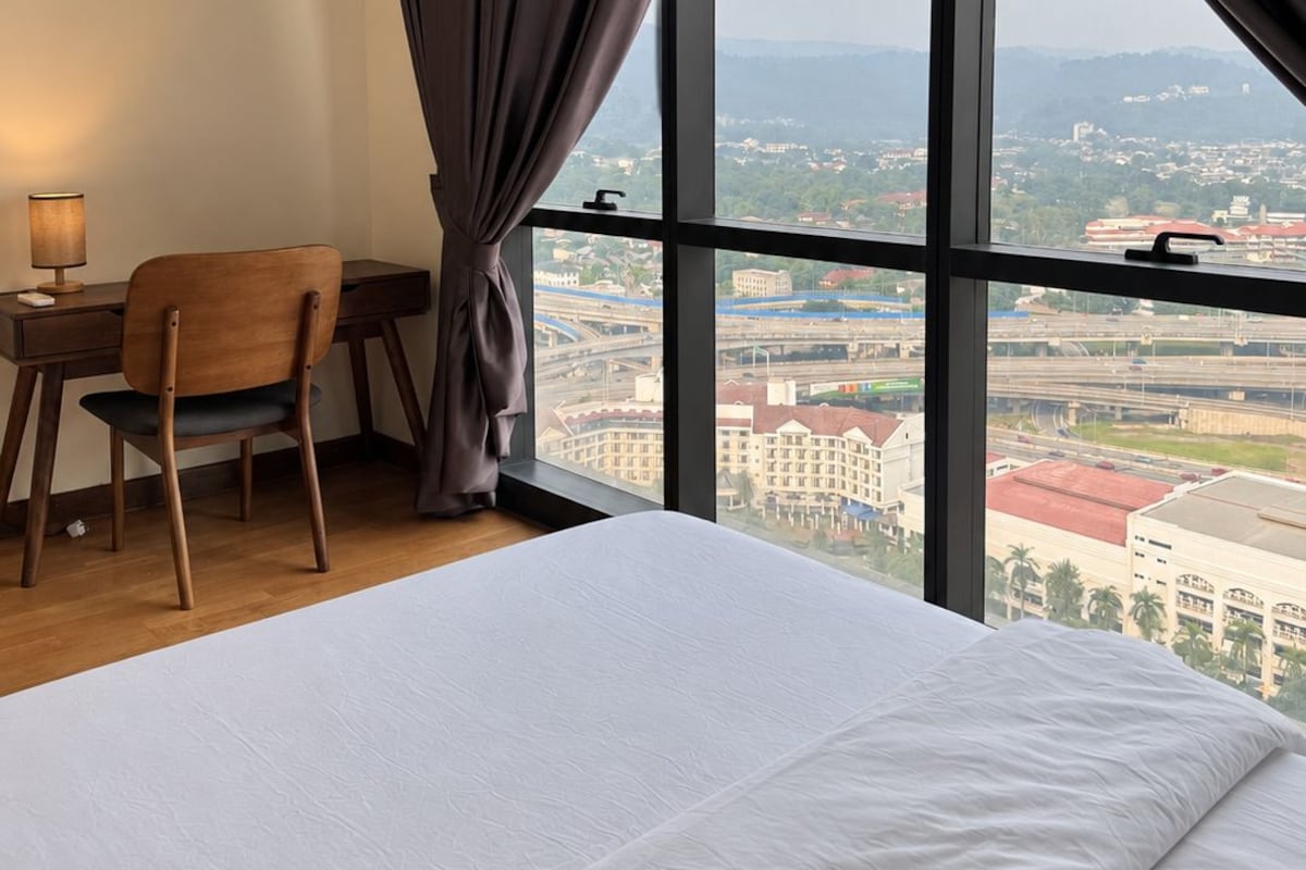 Ein Hotelzimmer mit einem großen Bett, einem Schreibtisch und einem Stuhl. Durch ein großes Fenster ist ein Blick auf die Stadt möglich.