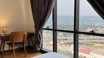 Ein Hotelzimmer mit einem großen Bett, einem Schreibtisch und einem Stuhl. Durch ein großes Fenster ist ein Blick auf die Stadt möglich.